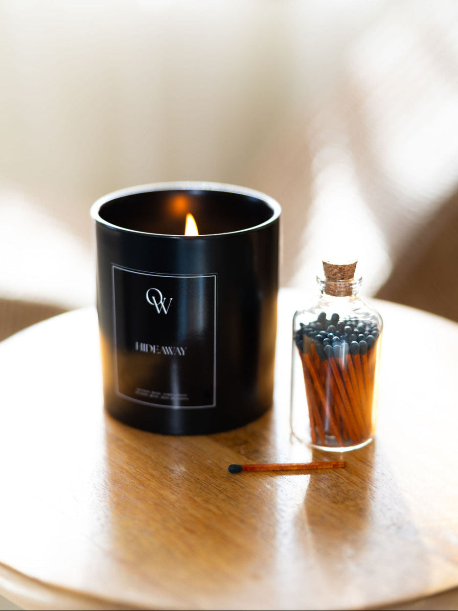 Black Hideaway candle beside a glass jar of black-tipped matches on a wooden table.